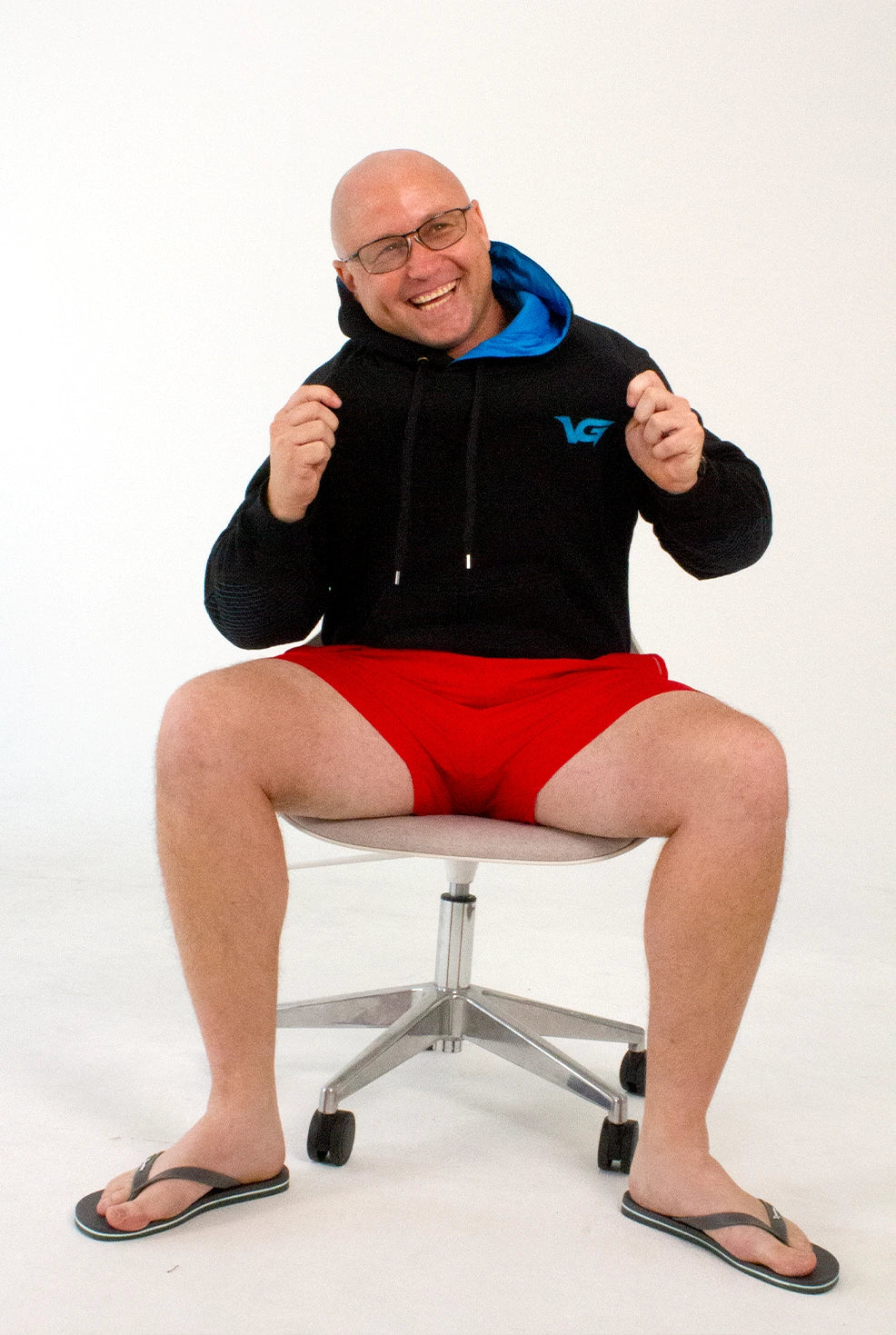 Man wearing a black hoodie with a logo and red shorts sitting on a chair against a white background